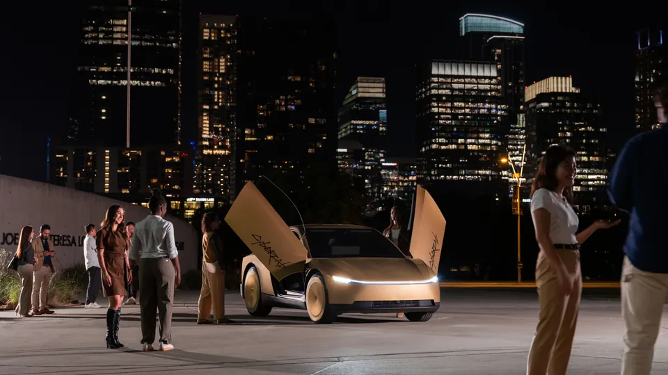 A gold Tesla Cybercab with both doors raised, parked at night in front of the Austin skyline as people stand around it