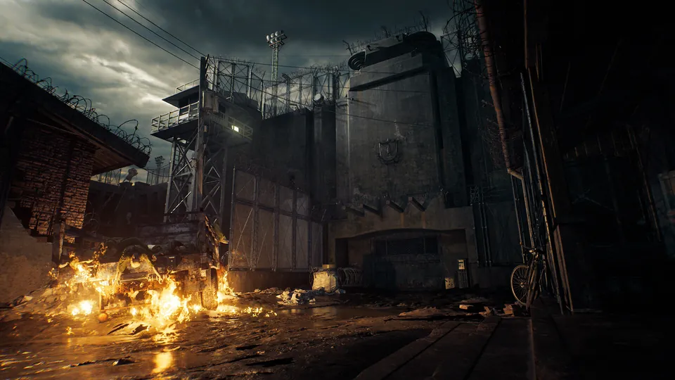 A burning wreck lights a muddy prison yard below a fortified gate, watchtowers and barbed wire under a stormy sky in Resident Evil Veronica