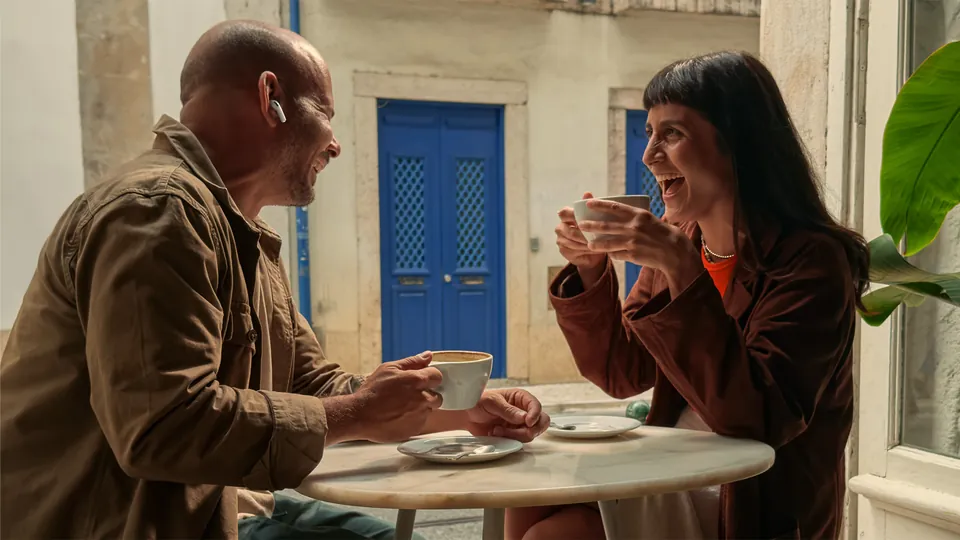 Two people talk over coffee, one wearing AirPods Pro 3