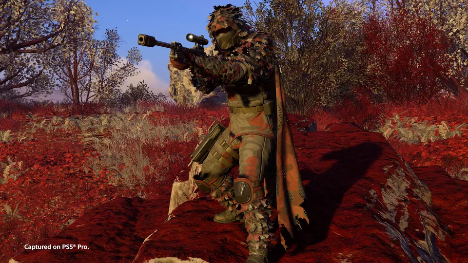 A Helldiver in ghillie camouflage aiming a scoped rifle in a field of red grass