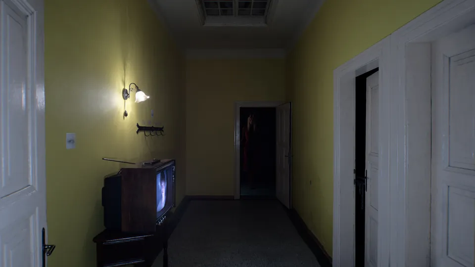 A dim yellow hallway with an old television showing a face and a figure in red standing in a far doorway, from OD
