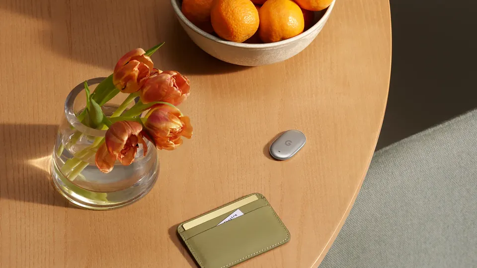 A small oval silver Pixel Tag sits on a wooden table beside an olive green cardholder wallet, a vase of orange tulips and a bowl of oranges