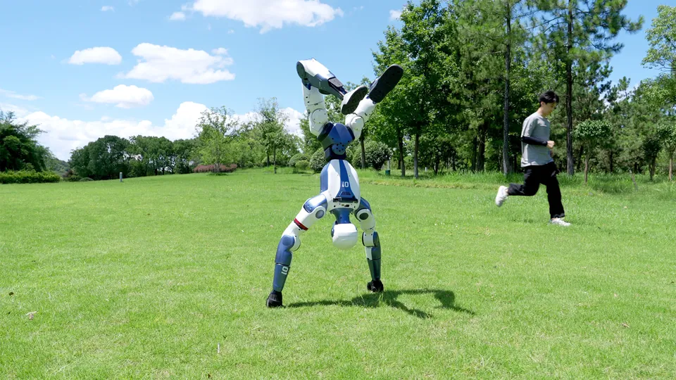 A white and blue Unitree R1 does a handstand on a lawn while a man jogs past