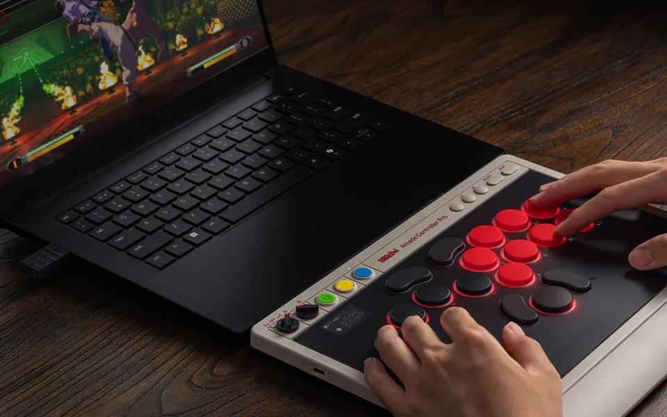 Two hands on the buttons of the Arcade Controller Pro N Edition beside a laptop running a fighting game, with the 8BitDo 2.4 GHz adapter plugged in