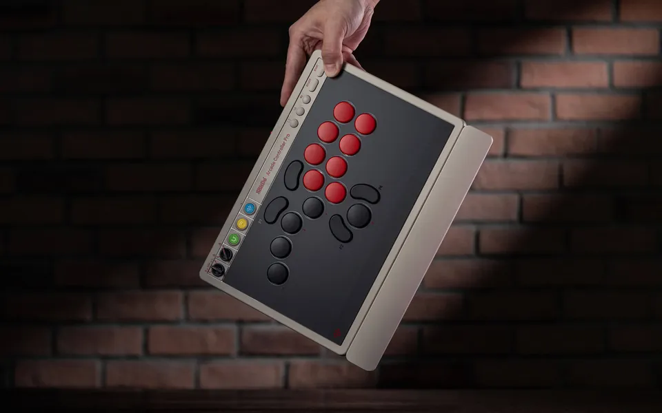 A hand holds the N Edition of the 8BitDo Arcade Controller Pro by one corner against a brick wall, with the wrist rest attached
