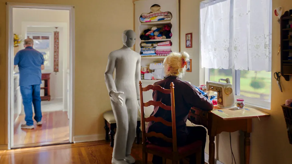 NEO stands beside an older woman sewing at a desk