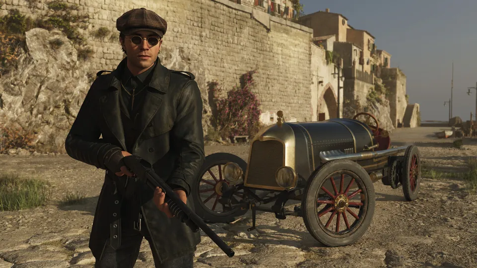 A man in a flat cap, round sunglasses and a long leather coat holds a shotgun beside a vintage race car on a sunlit Sicilian street in Mafia: The Old Country