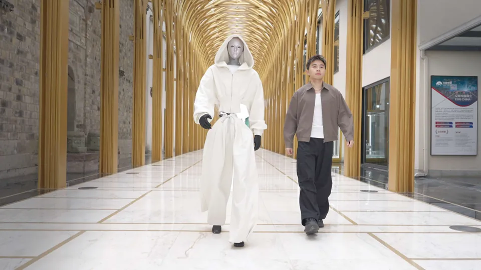 The Unitree H2 in a white hooded outfit walking beside a man down a hall lined with golden arches