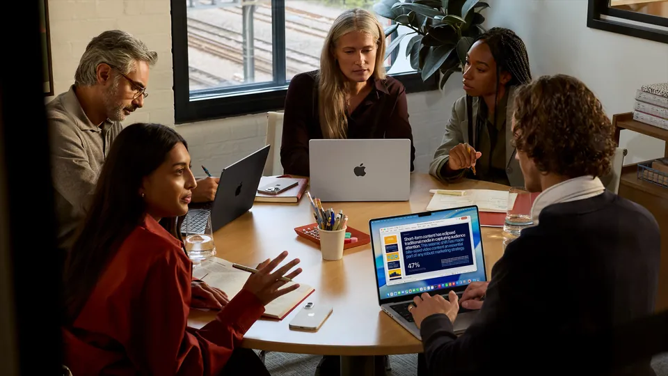 Five coworkers around a meeting table with MacBook Pro laptops