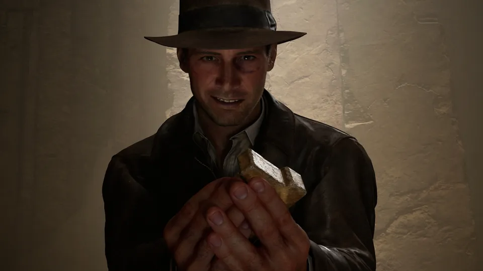 Indiana Jones in a fedora and leather jacket smiles while holding a small stone artifact in a dim room