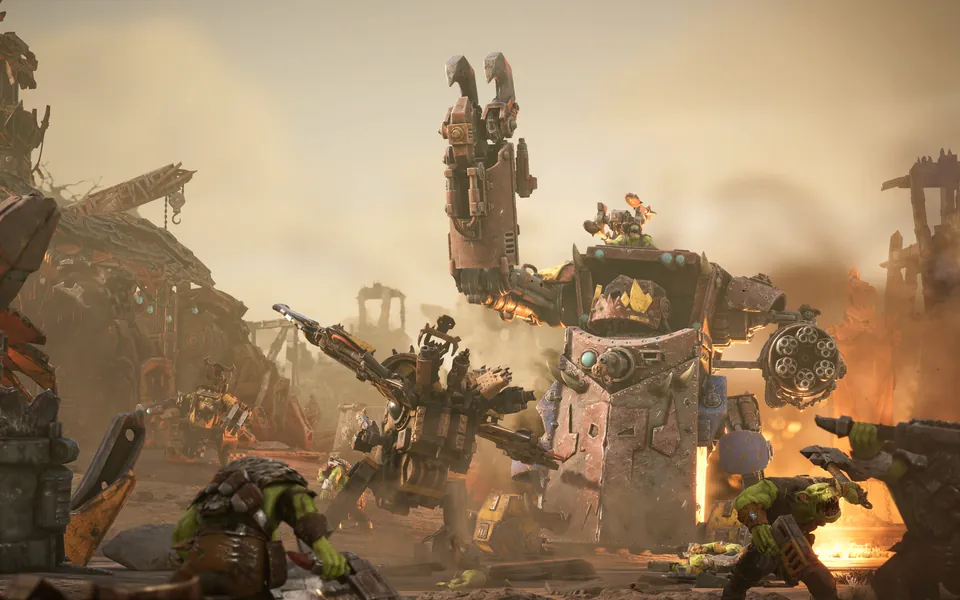 A huge Ork war machine with a claw arm and a rotary gun stomps through a burning scrapyard as Ork warriors fire guns in Dawn of War IV