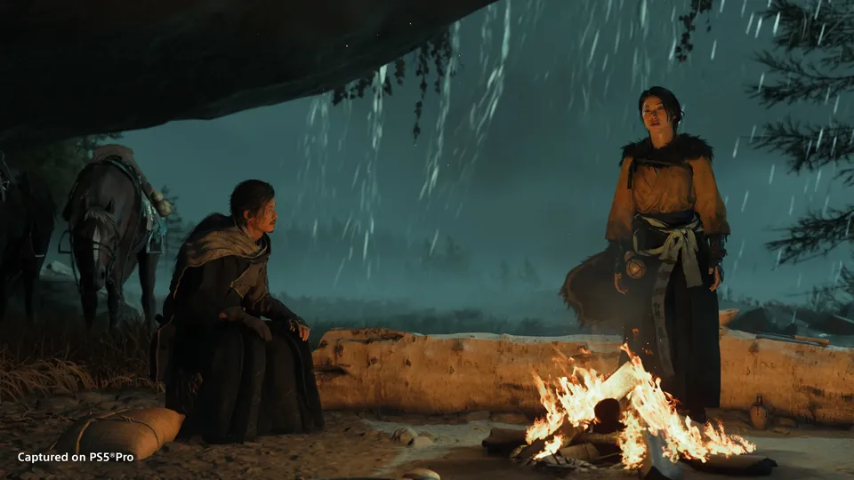 Atsu stands by a campfire under a rocky overhang in the rain while a companion sits nearby in Ghost of Yōtei