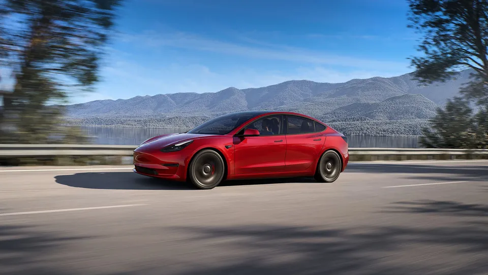 A red Tesla Model 3 driving along a lakeside road with mountains behind