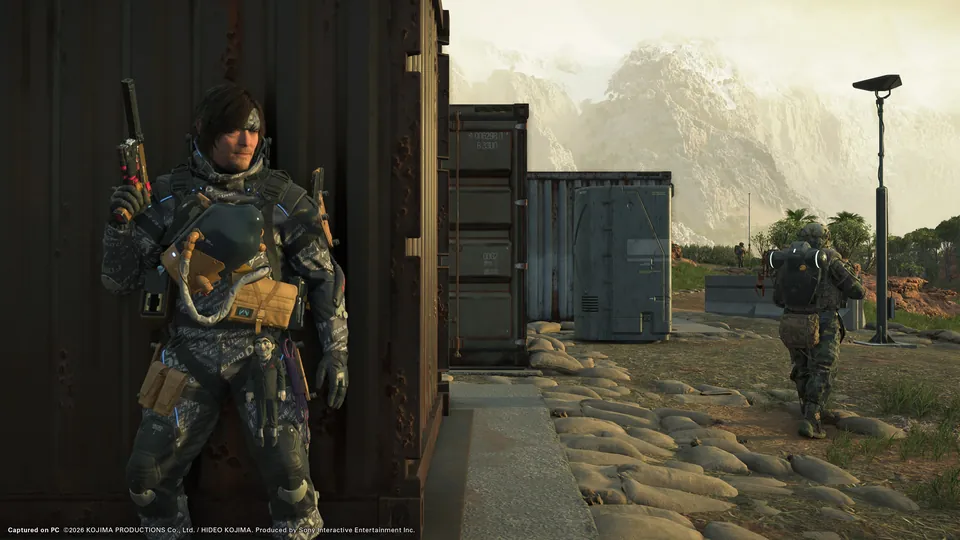 Sam Porter Bridges leans against a rusty shipping container holding a pistol at a guarded camp below snowy mountains in Death Stranding 2: On the Beach