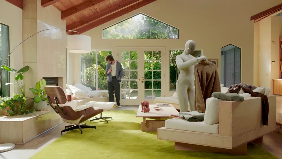 The NEO humanoid robot tidies a living room while a person walks in from the garden