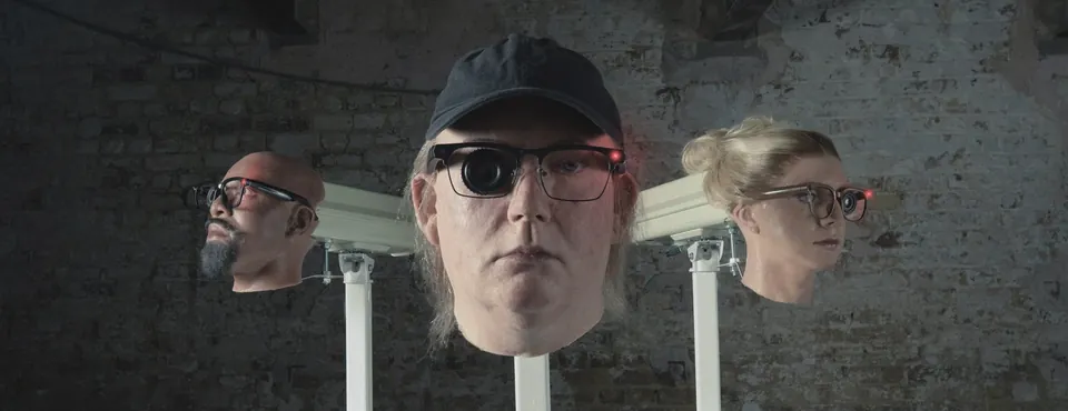 Three lifelike heads mounted on poles like security cameras, each wearing glasses with a camera