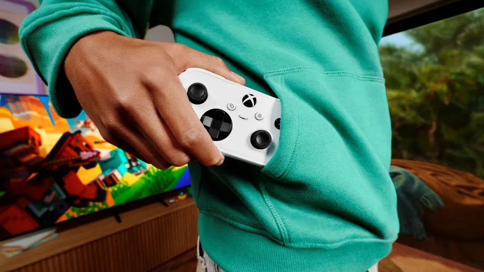 A small white Xbox Nano Controller held in one hand in front of a TV
