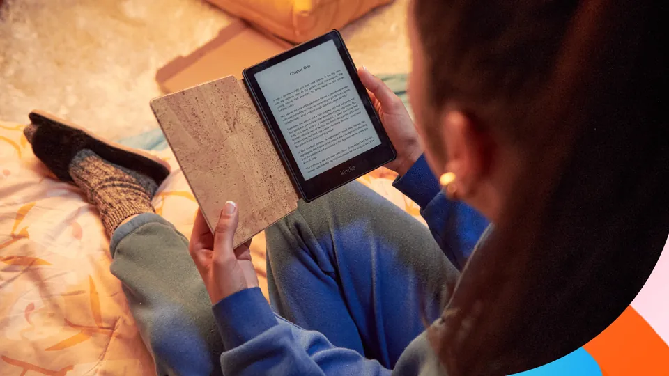 A reader in a blue sweater holds a Kindle e-reader in a cork cover while sitting on a bed
