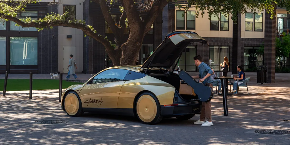 A man loading a guitar case into the open rear hatch of a gold Tesla Cybercab parked on a city plaza