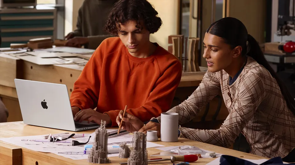 Two people work at a wooden workbench with a silver 14-inch MacBook Pro and architectural models