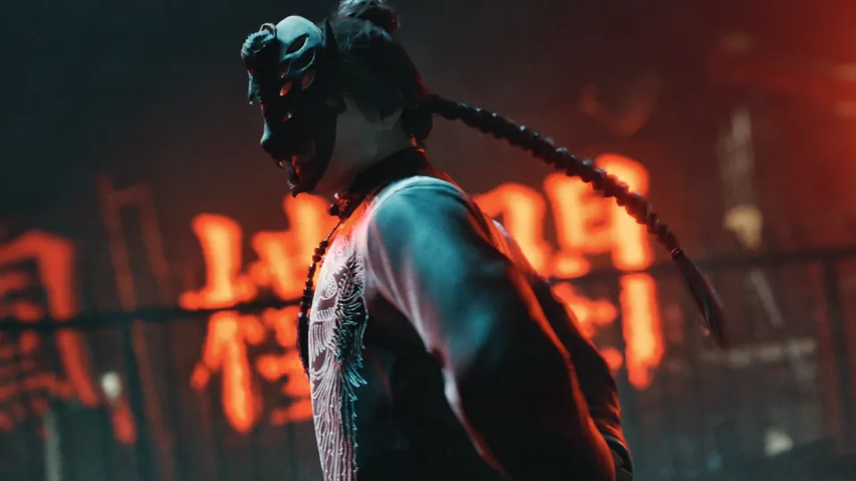 A masked figure with a long braid stands in front of glowing red neon characters at night