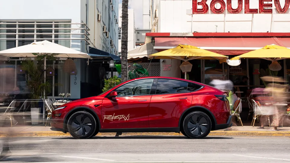 A red Tesla Model Y with Robotaxi lettering parked on a street in front of a cafe with yellow umbrellas