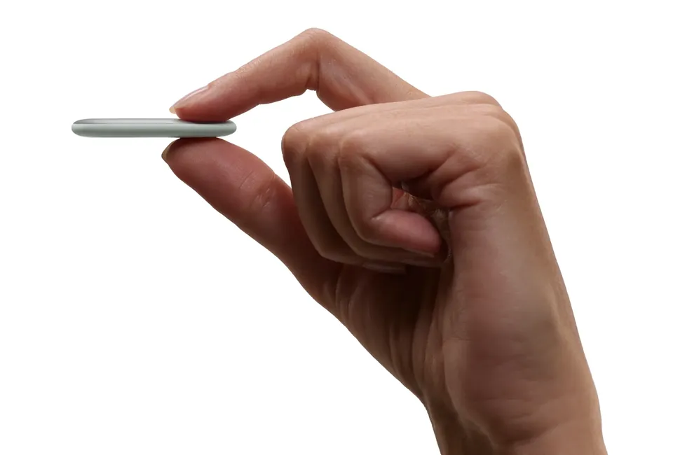 A hand holds the thin, pale gray Pixel Tag edge-on between thumb and finger against a white background