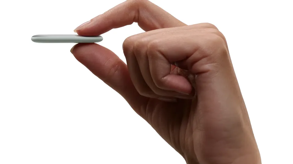 A hand holds the thin, pale gray Pixel Tag edge-on between thumb and finger against a white background