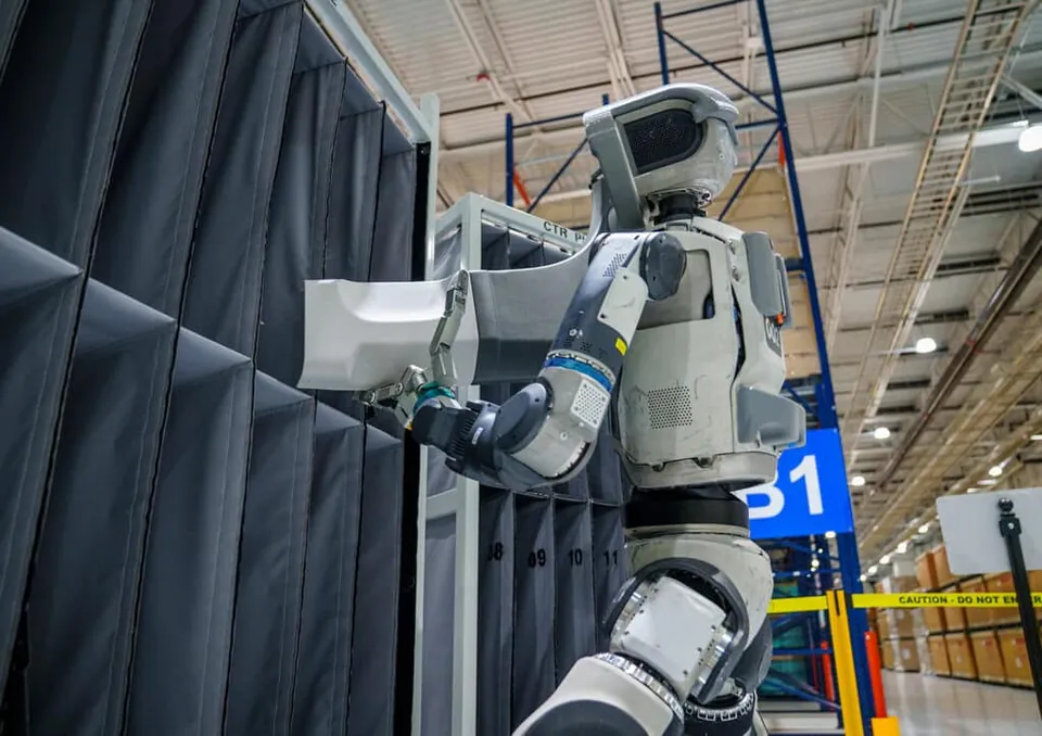 Atlas reaches into a parts bin in a factory aisle