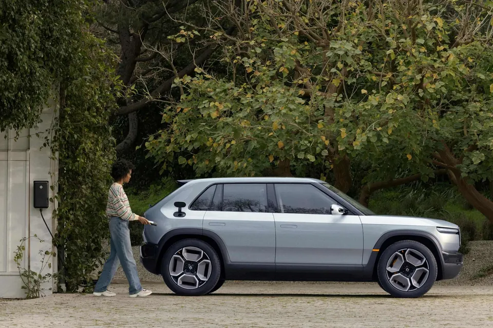 A person walking past a Rivian R3 parked in a driveway next to a wall-mounted charger
