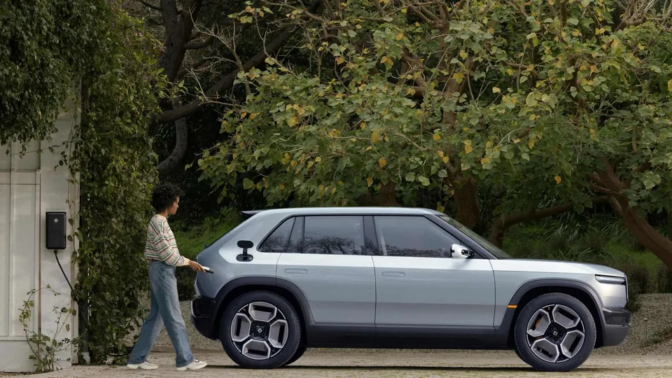 A person walking past a Rivian R3 parked in a driveway next to a wall-mounted charger
