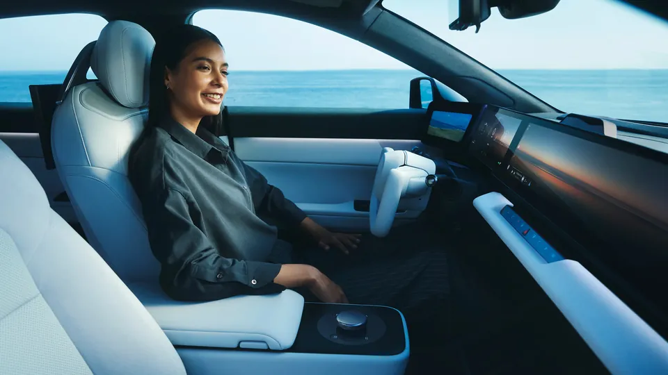 A woman in the driver's seat of an Afeela 1, with a yoke-style steering wheel and a wide screen across the dashboard