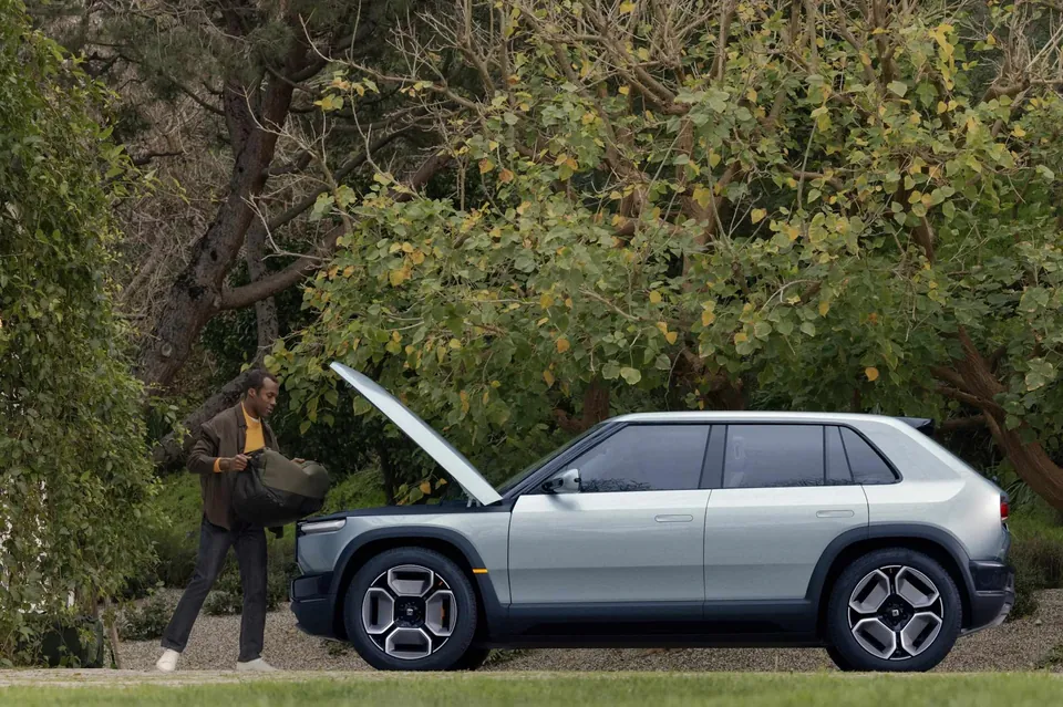 A person lifting a bag from the open front trunk of a Rivian R3 parked under trees