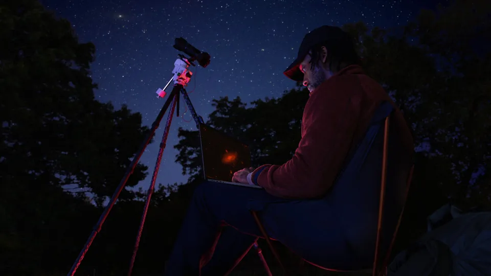 A stargazer uses a MacBook Pro next to a telescope at night